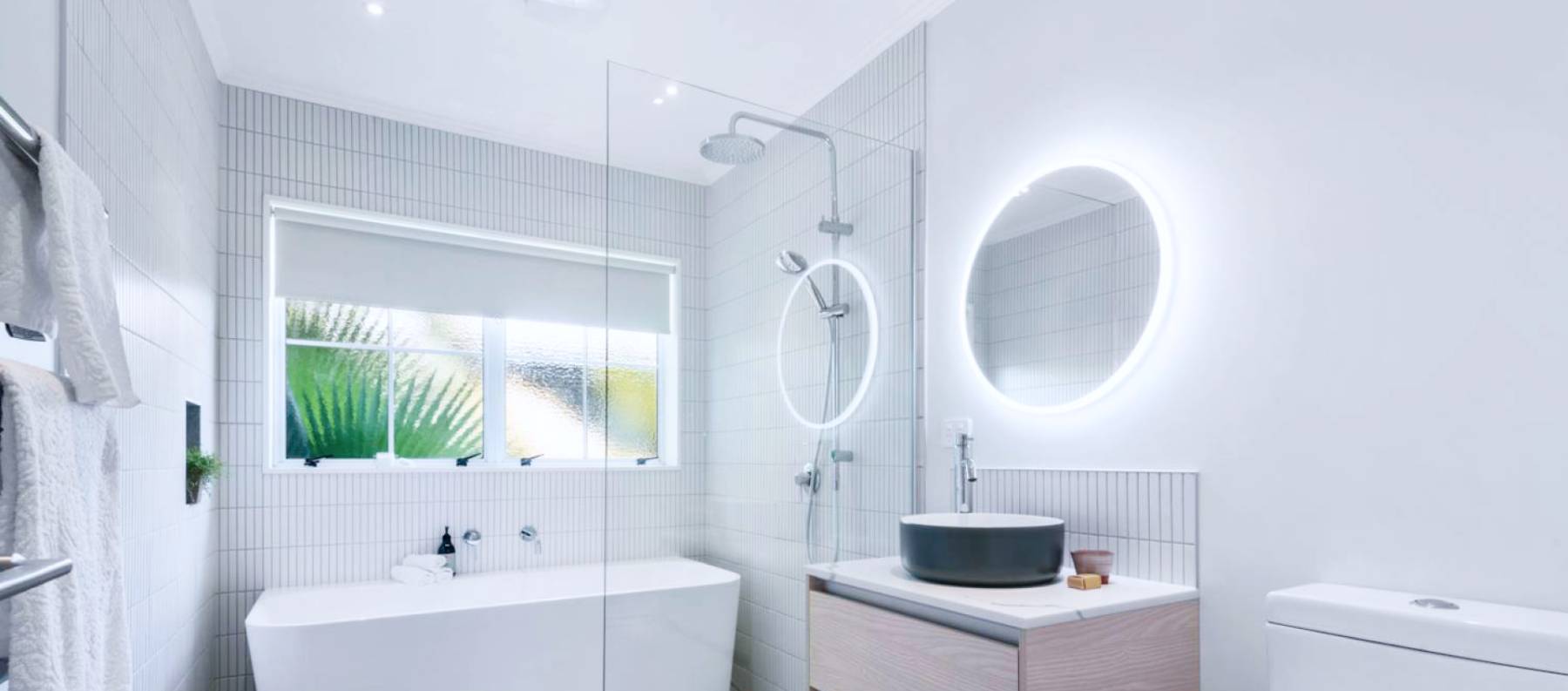 Renovated bathroom with tiled bath and separate rain shower area, glass screen, vanity and circular mirror with LED light around